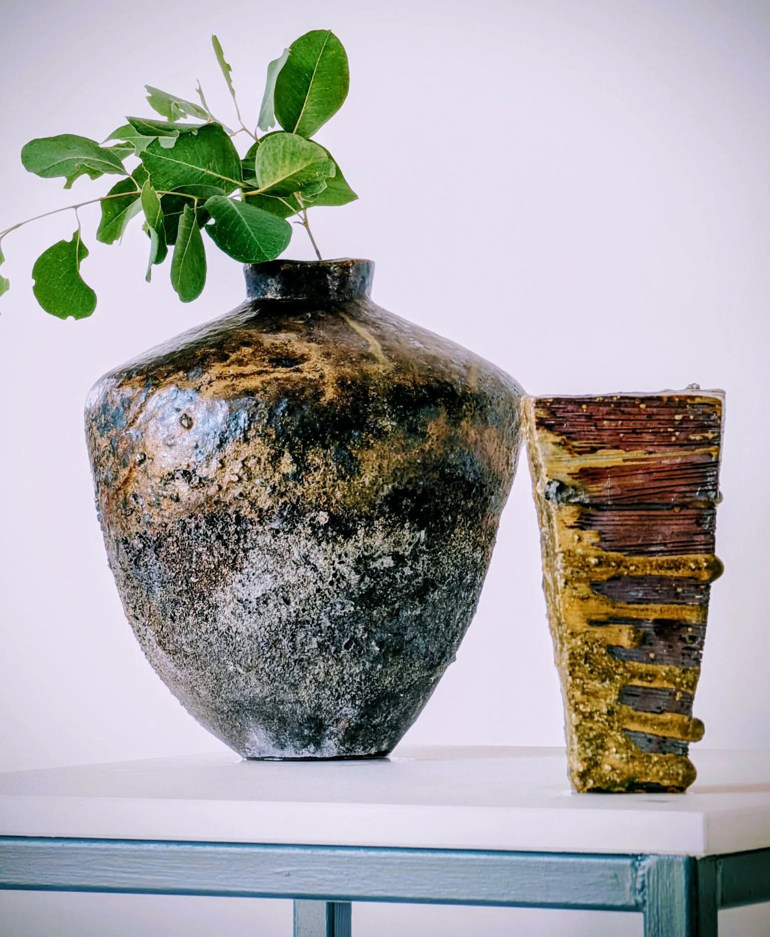 A large, rustic ceramic vase with a black and brown marbled glaze, holding a green leafy branch. To the right of the vase is a textured wooden sculpture with horizontal lines, sitting on a white surface against a plain light background.