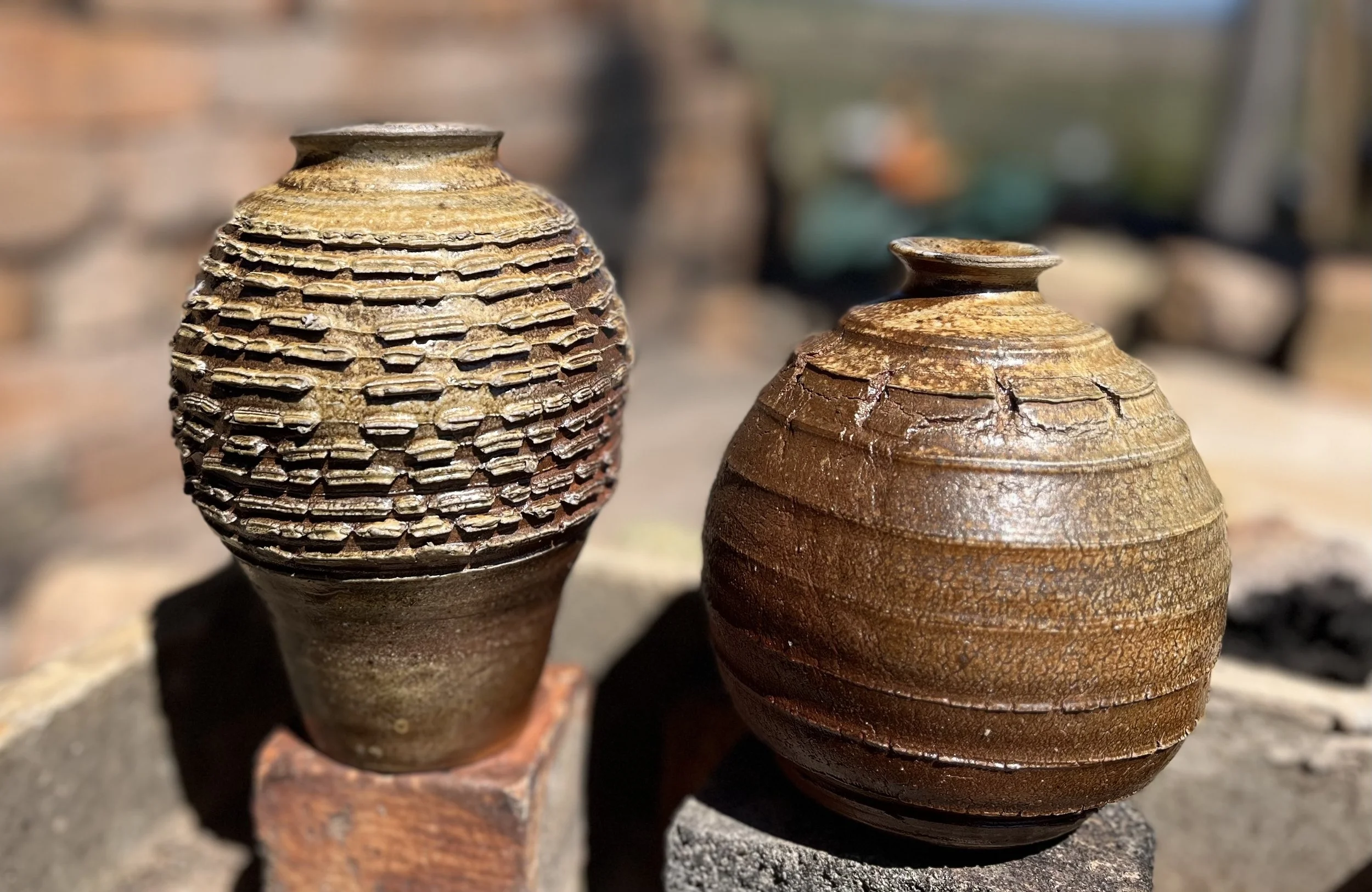 Two ceramic vases with textured surfaces are displayed outdoors on stone supports, with the anagram kiln at Gundaroo in the background.