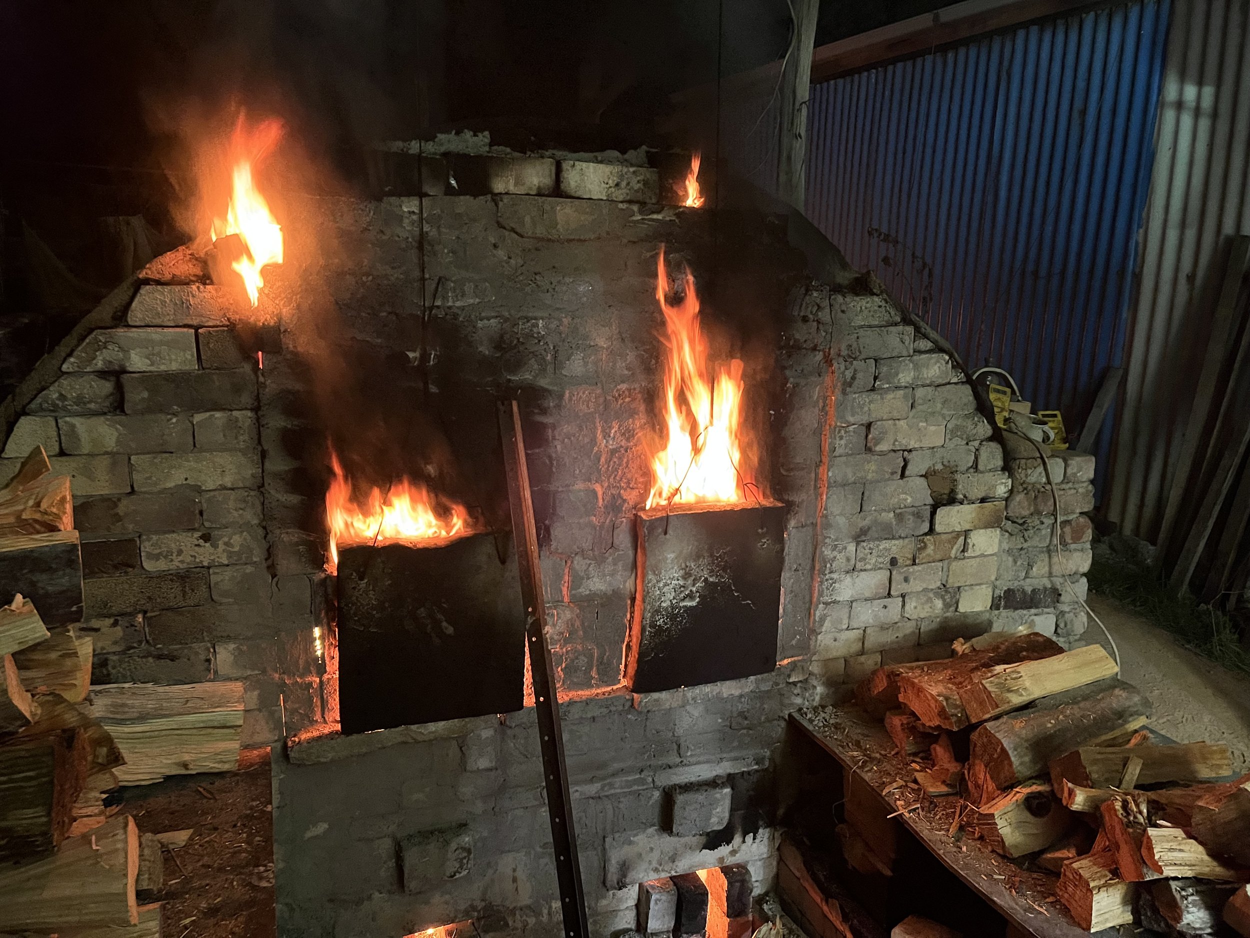 Fire burning in a brick oven or fireplace with stacked wood to the side.
