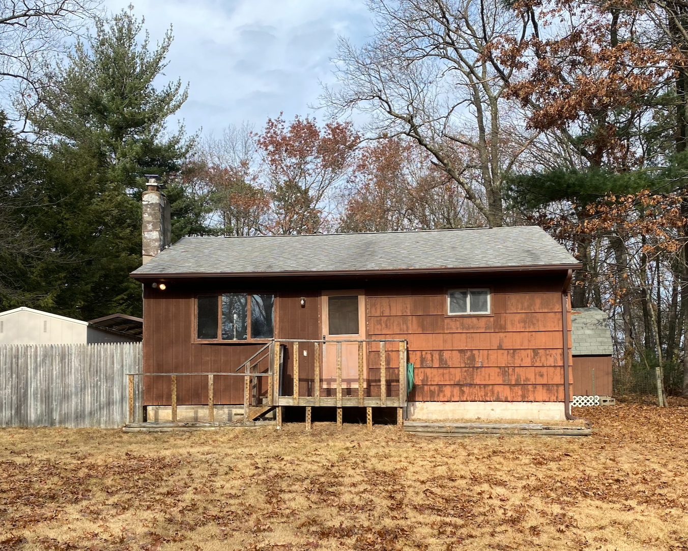Back of a small wooden house with a porch, surrounded by trees with fall foliage, and a grassy yard in the foreground.