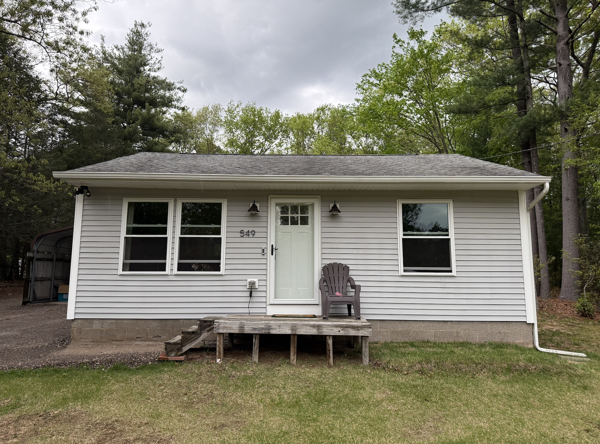 Front of a small house with white siding, two windows on each side of a white door, and a wooden staircase with three steps. A purple lawn chair with a pink object on it is on the porch. The house has two outdoor lights above the door, and the yard has grass and trees in the background.