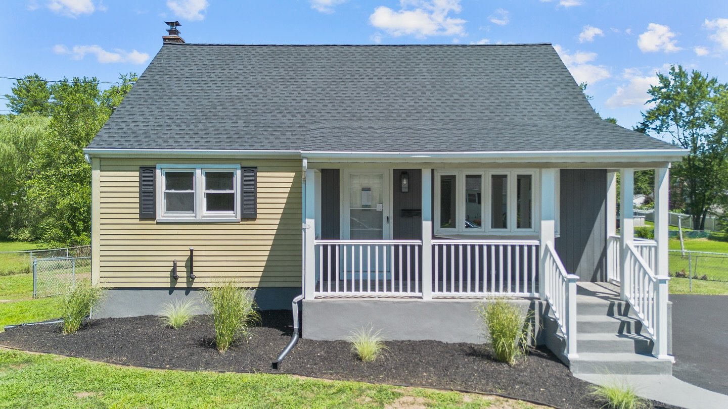 Front view of a newly built house with a porch, stairs, a gable roof, beige and gray siding, black shutters, and a small yard with bushes and grass, under a blue sky with clouds.