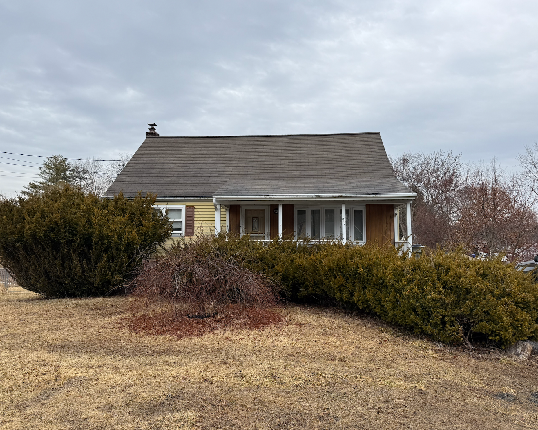 A house with yellow siding and brown shutters, surrounded by bushes and grass, under a cloudy sky.