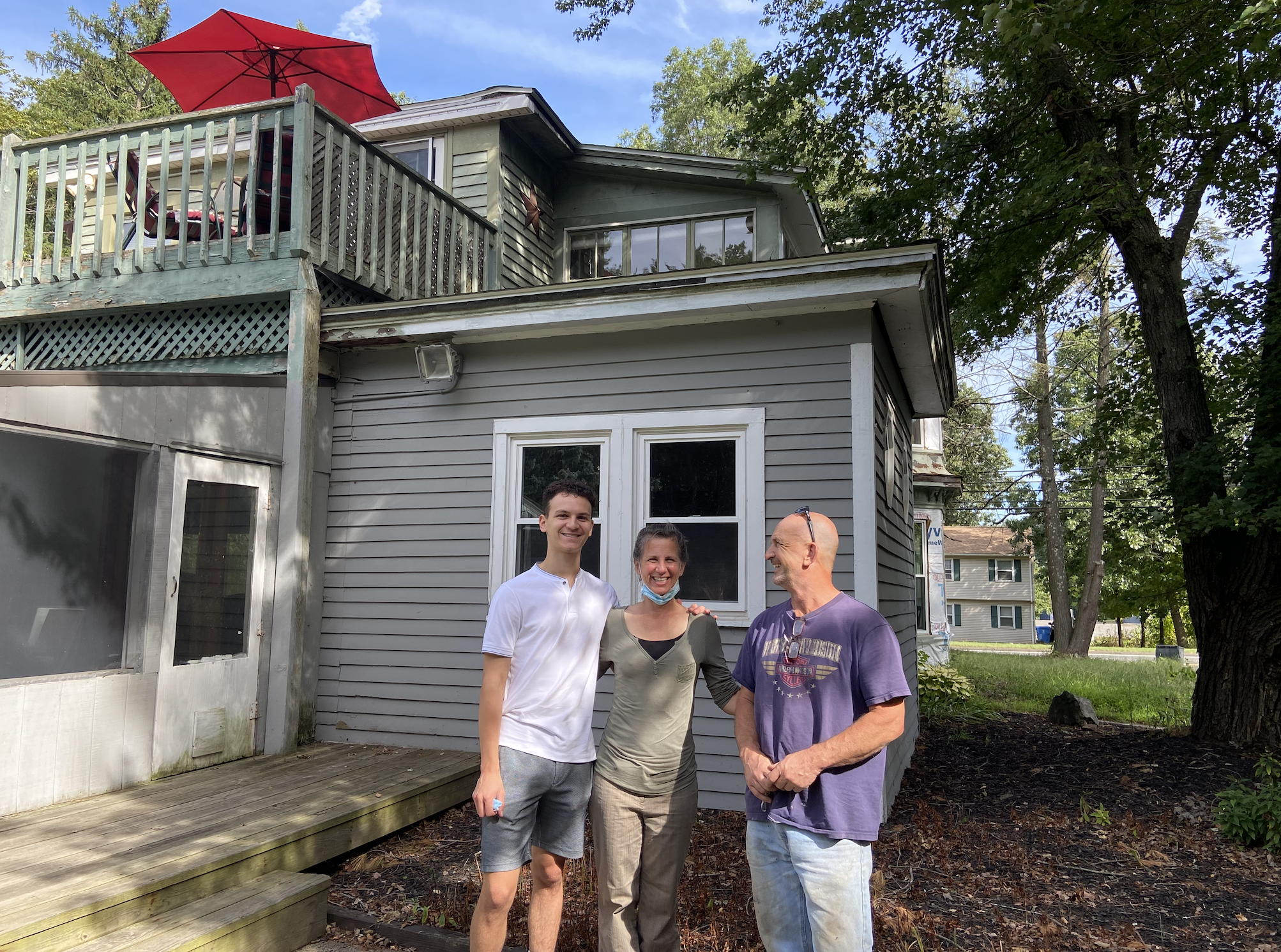 Three people smiling and standing together outside a house with gray siding and a wooden deck. One person has an arm around another, and they are all facing the camera. The scene is in a yard with trees and other houses in the background.