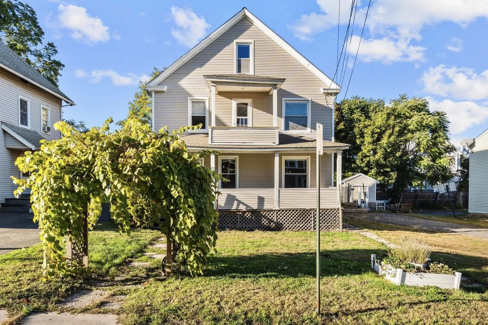 A three-story beige house with a front porch, balcony, and multiple windows, surrounded by trees, with a clear blue sky in the background.