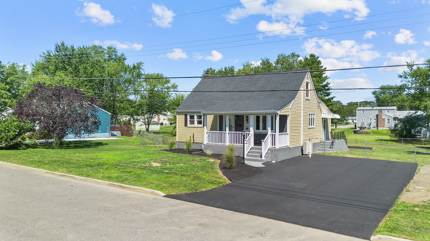A new yellow house with a black roof and white porch, located on a residential street. The driveway is freshly paved with black asphalt. Surrounding greenery and other houses can be seen in the background.