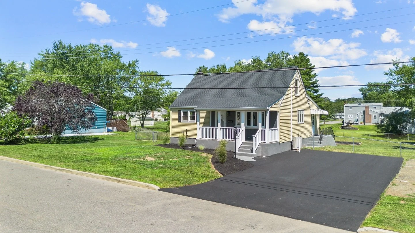 A new yellow house with a black roof and white porch, located on a residential street. The driveway is freshly paved with black asphalt. Surrounding greenery and other houses can be seen in the background.