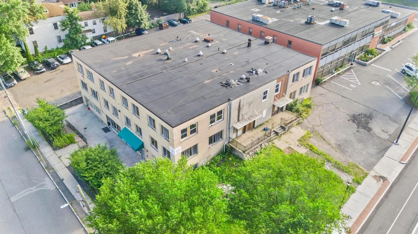 An aerial view of a three-story beige building with a flat roof, surrounded by green trees and a parking lot with a few cars. Part of a larger parking lot is visible on the right, and a street runs along the bottom of the image.