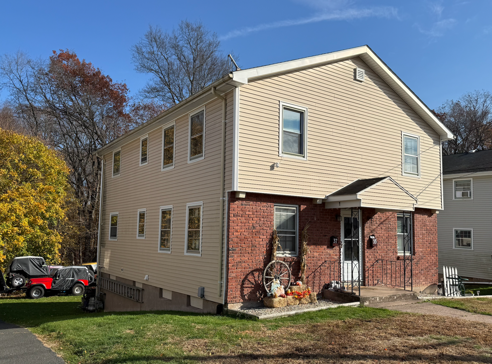A two-story house located in Connecticut, with beige siding and a brick front entrance, surrounded by a lawn and trees with fall foliage, under a blue sky.