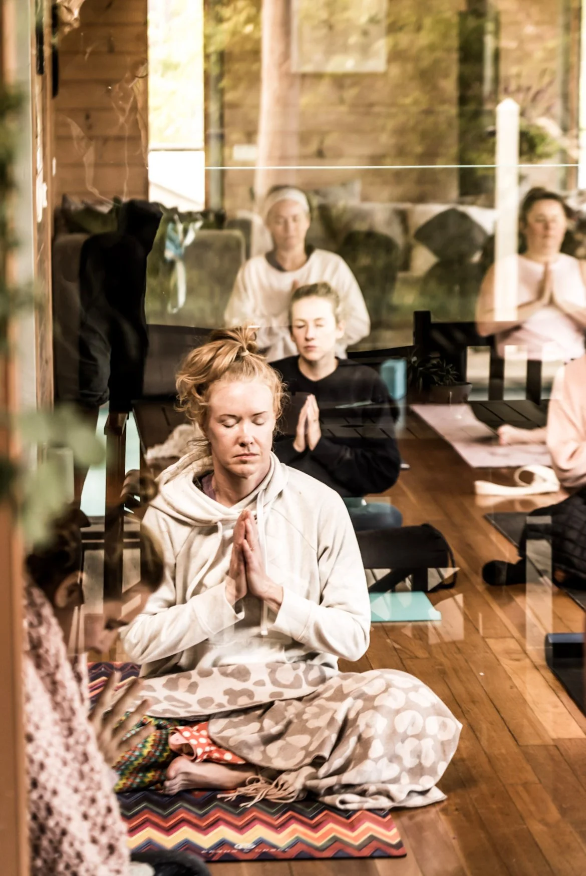 People practicing meditation in a peaceful indoor setting, sitting cross-legged with hands in prayer position, eyes closed, on colorful mats, behind a glass window reflecting trees and sunlight.