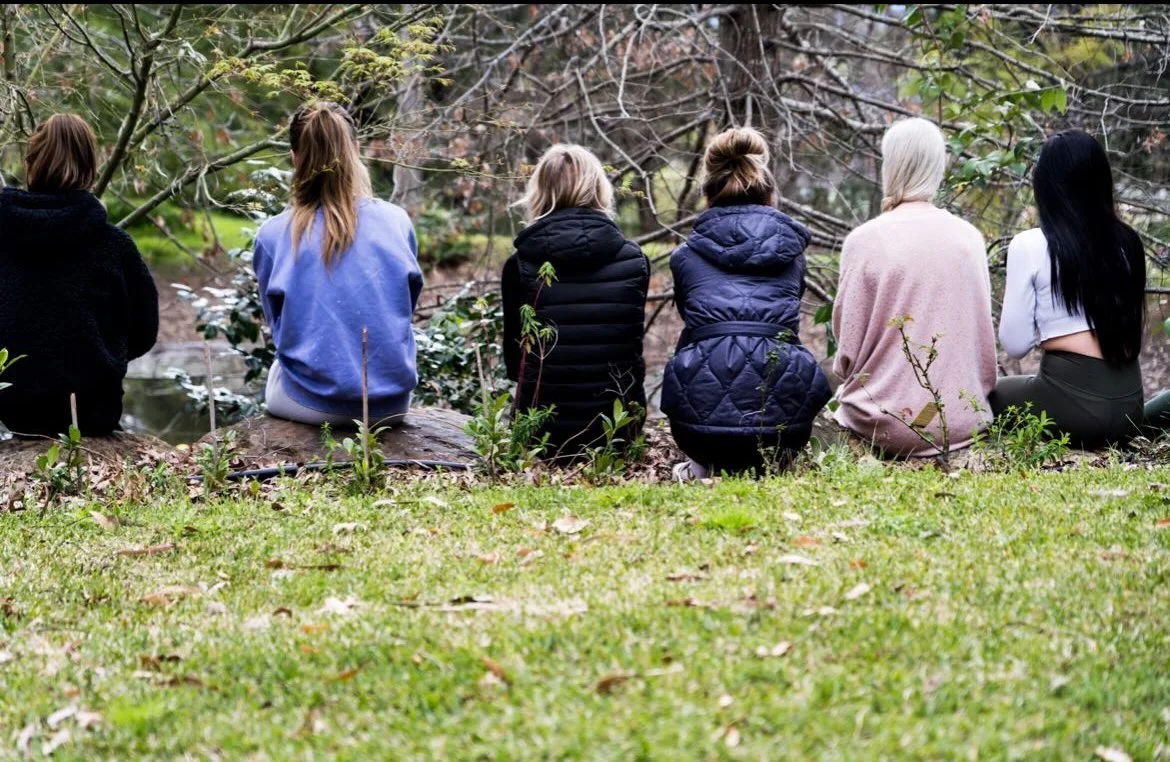 Six women sitting on logs by a pond, facing away in a wooded park.