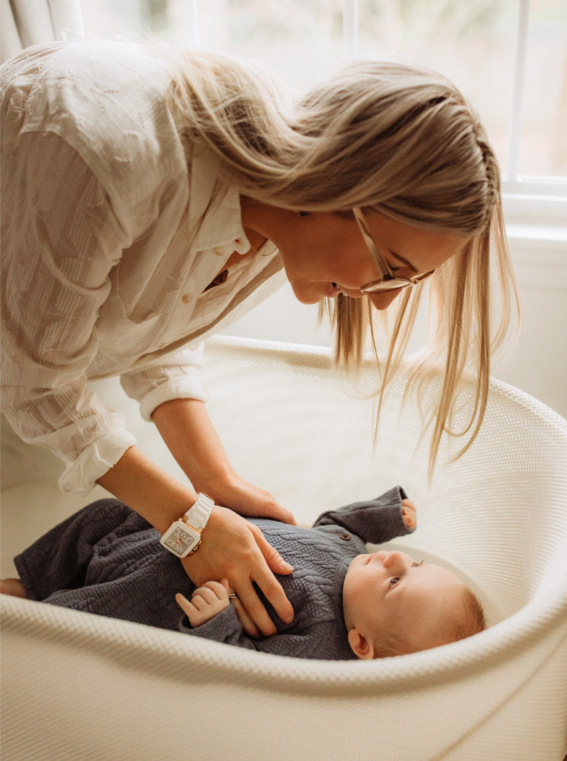 woman in white standing over a newborn in grey jumper lying in a bassinet