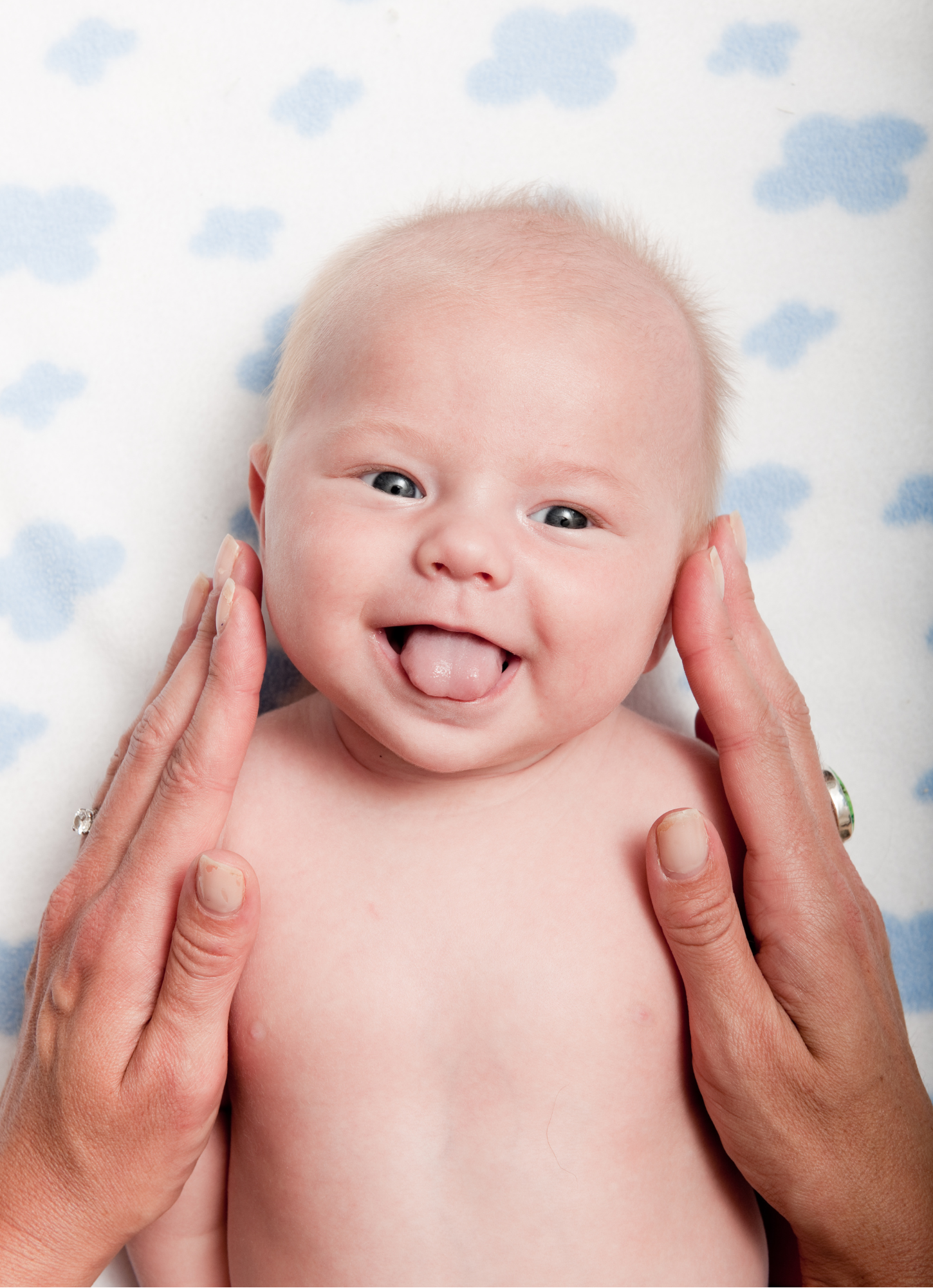 A smiling baby with blue eyes sticking out their tongue, held gently by two adult hands, lying on a blanket with blue clouds pattern.
