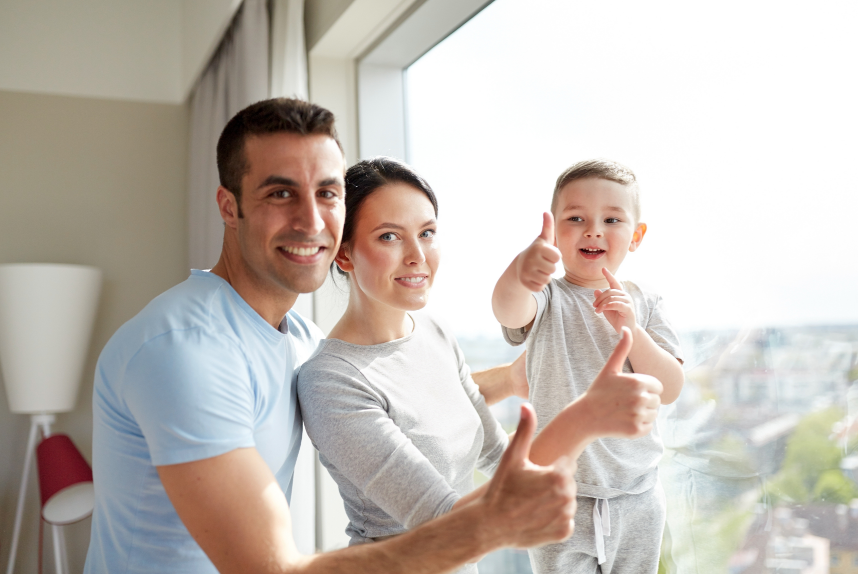 A happy family of three, a father, mother, and young son, standing together by a large window giving thumbs up and smiling.