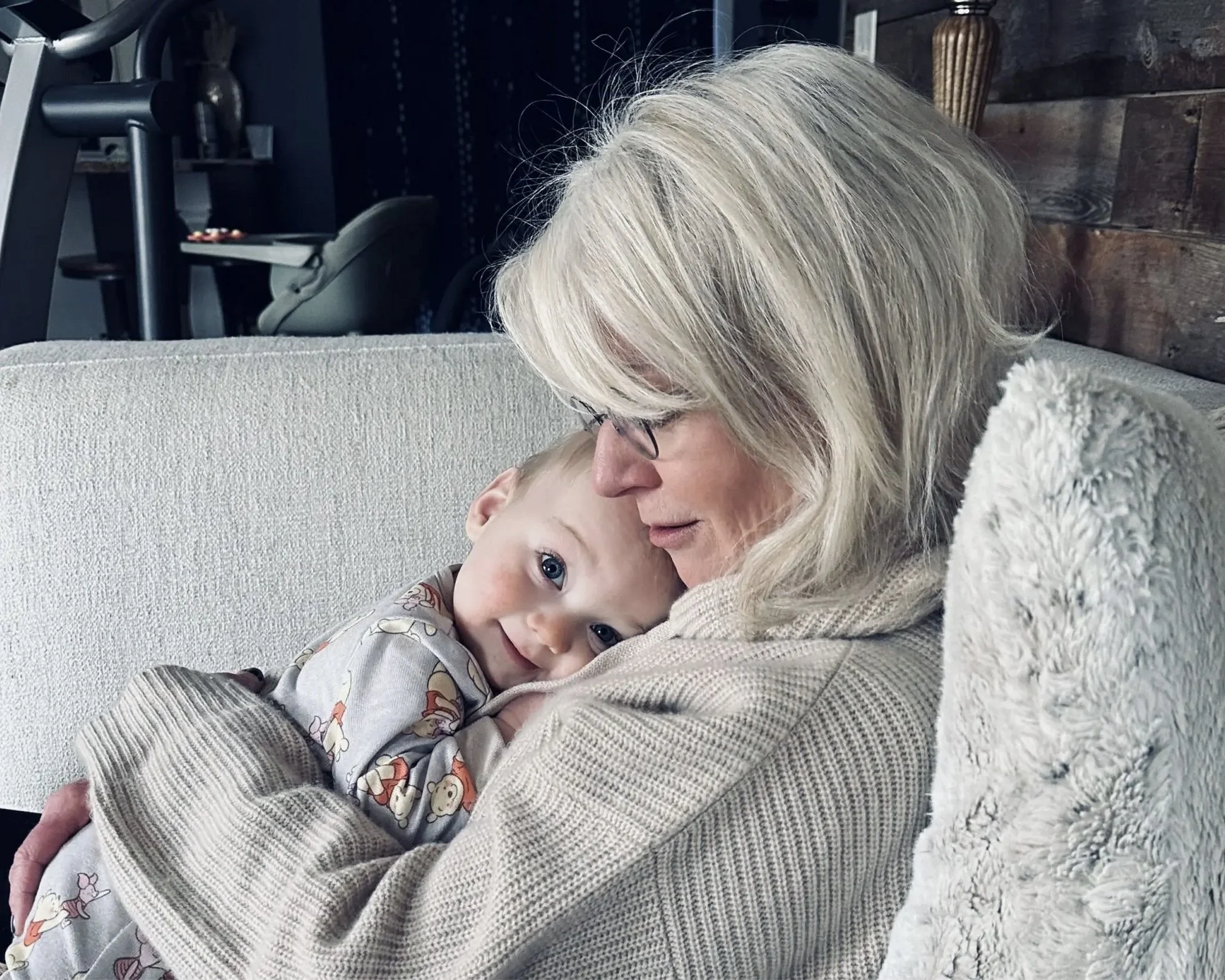 An older woman with blonde hair and glasses cuddling a young child with blonde hair and blue eyes, sitting on a light-colored sofa in a cozy indoor setting.