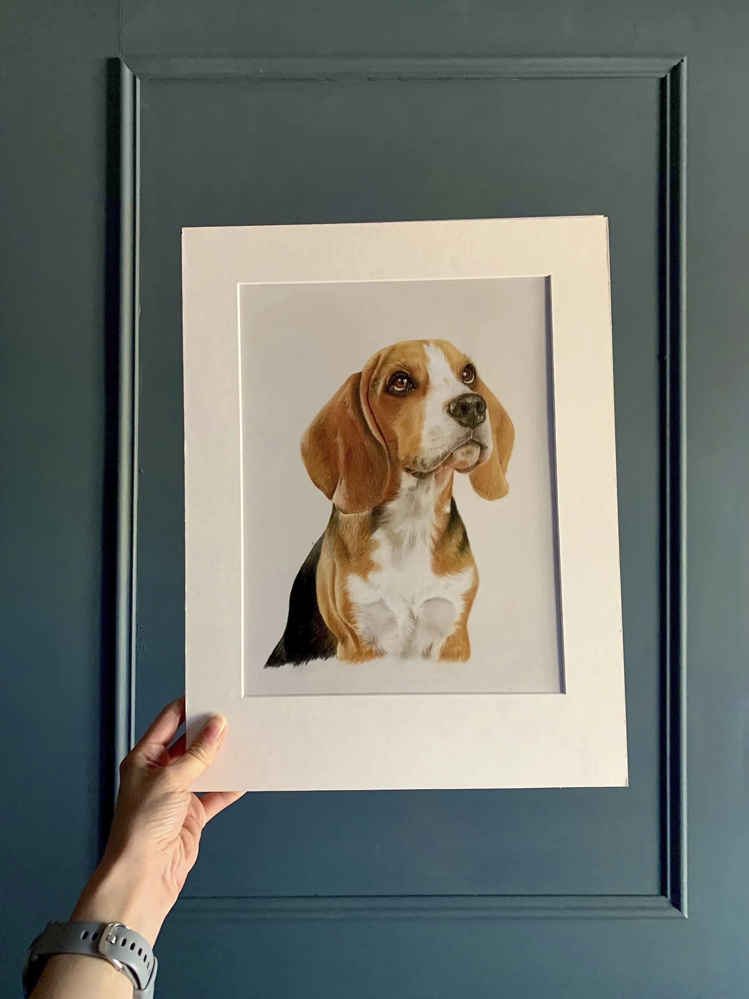Hand holding a framed portrait of a beagle dog in front of a teal door.