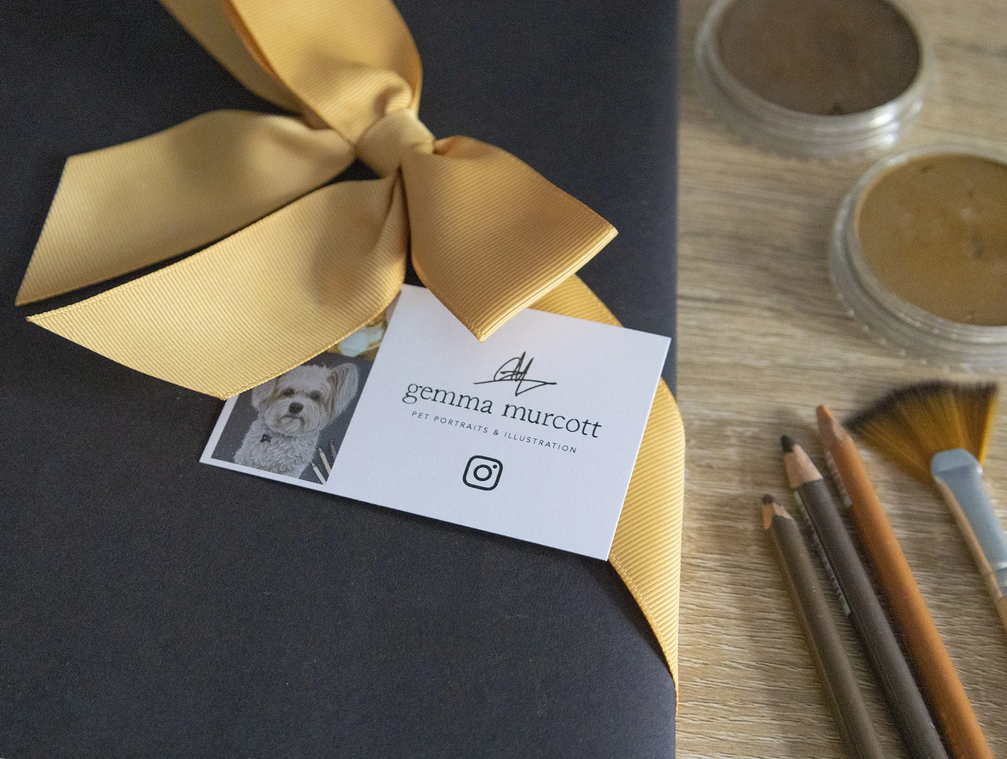 A business card for Gemma Murcott with a pet portrait photo, black text, and Instagram icon, placed on a black surface with a large yellow bow, alongside pastel pencils on a wooden surface.