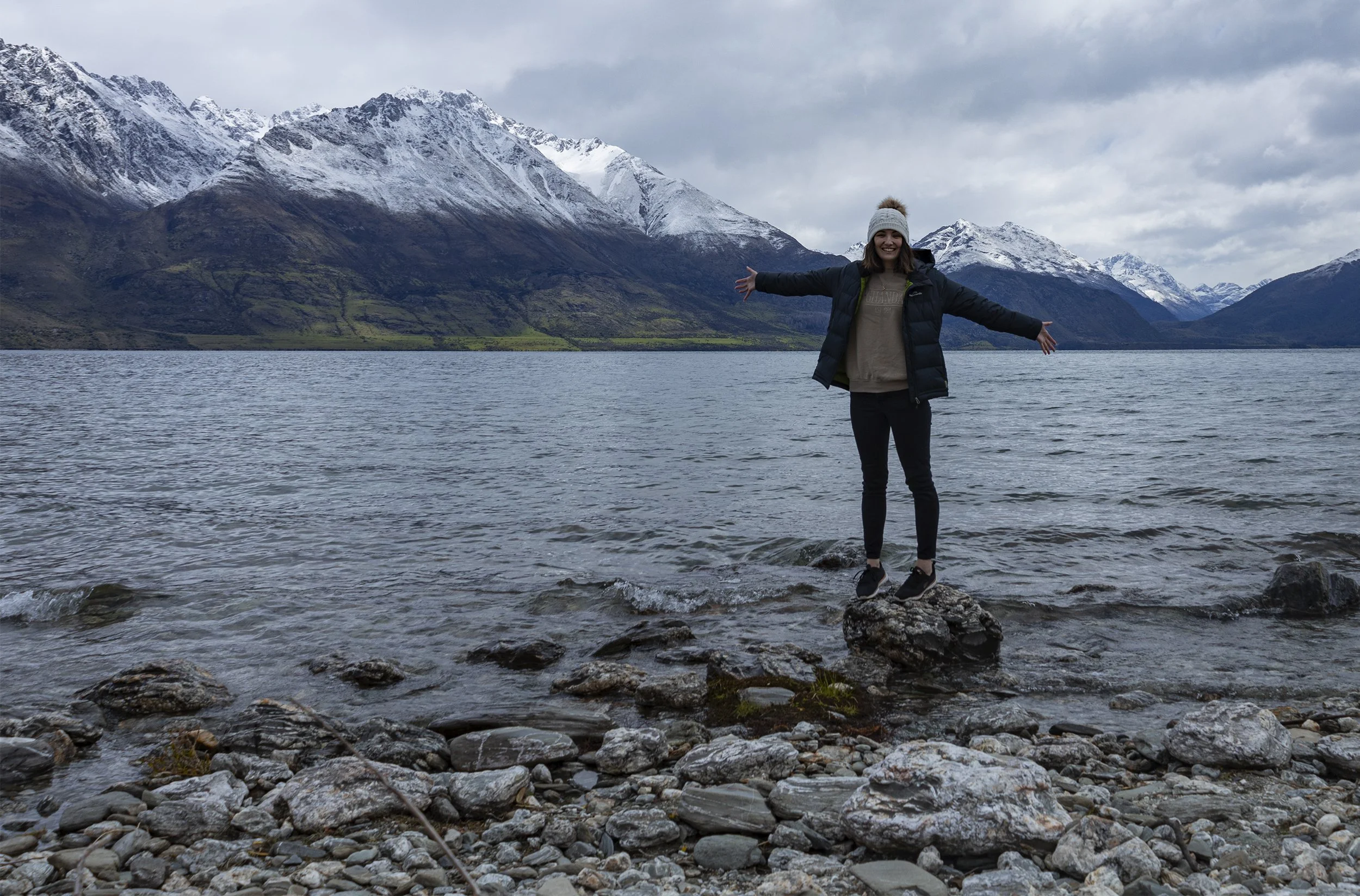 A woman standing on a rock by a lake with snow-capped mountains in the background, wearing a winter jacket and beanie, arms outstretched.
