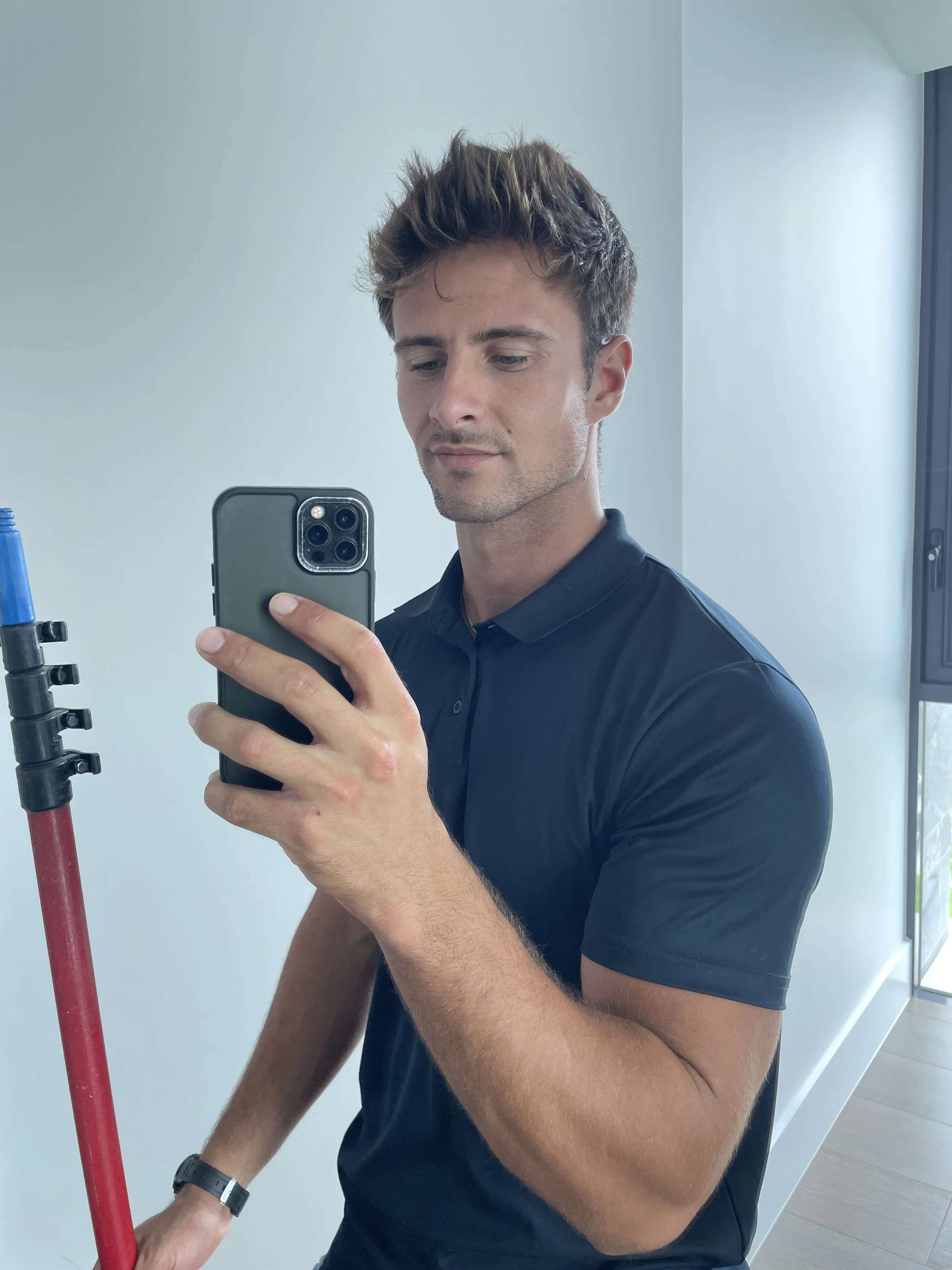Man taking a selfie with a smartphone while holding a red pole, wearing a black polo shirt.