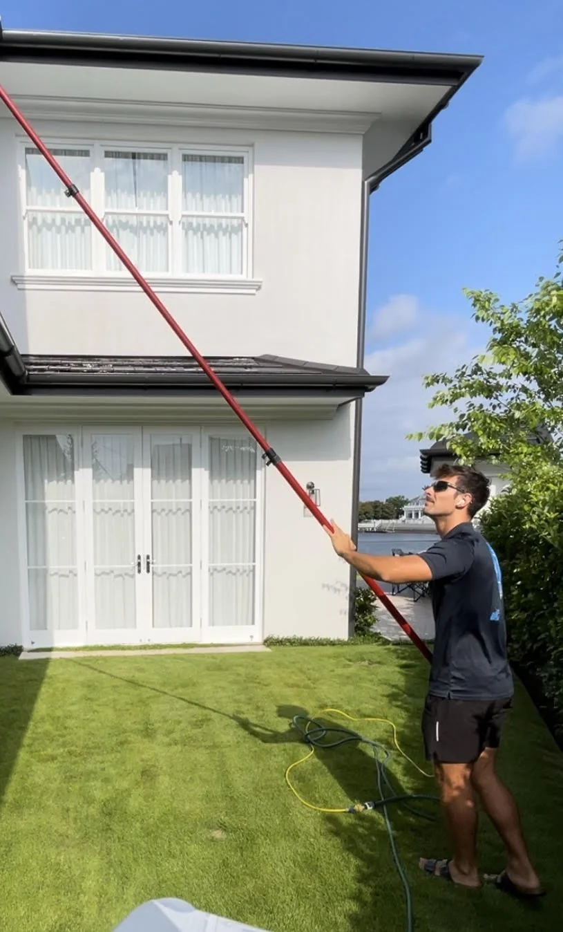Person cleaning second-story windows with a long pole on a grassy lawn.