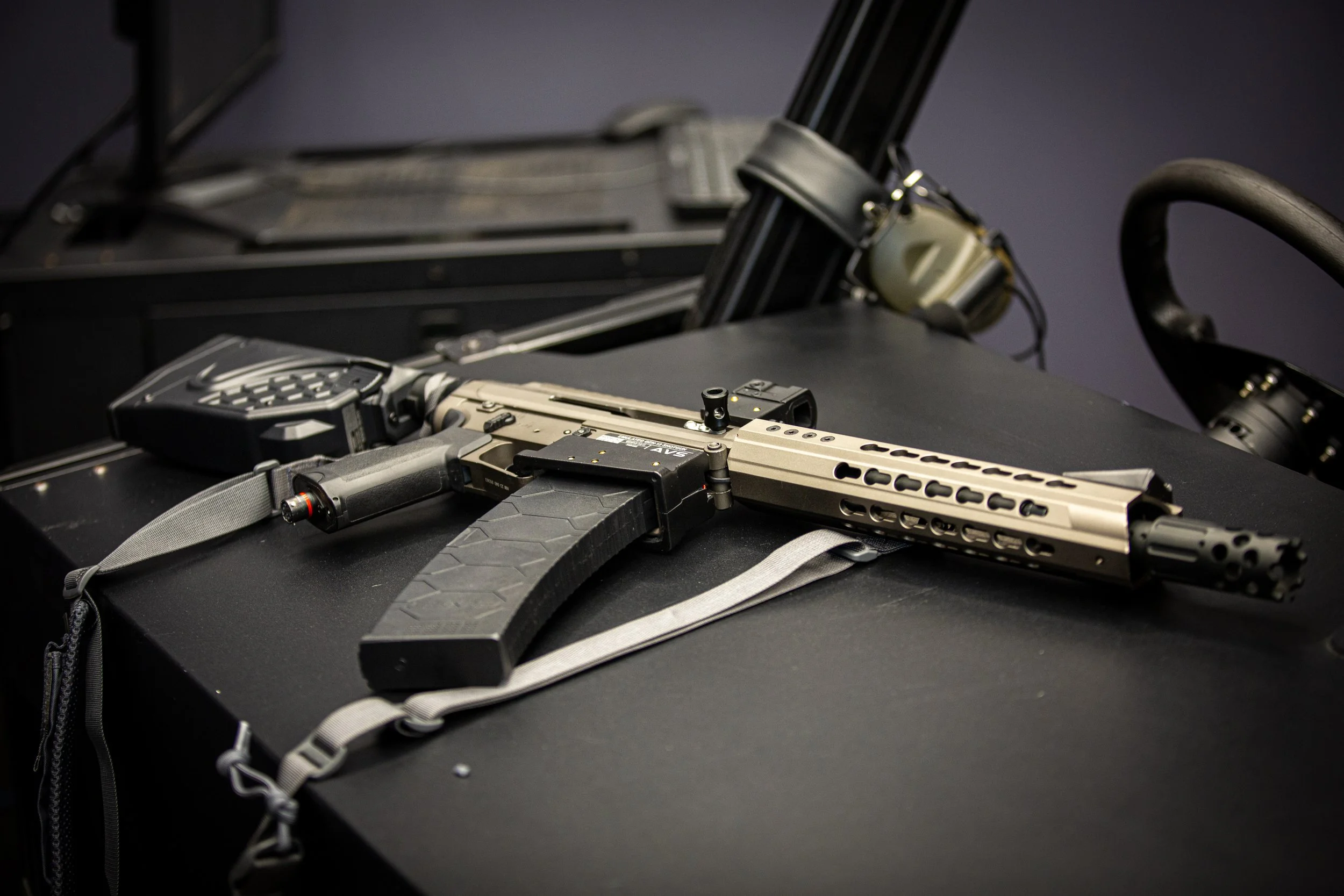 A firearm with a tan and black color scheme, included with accessories, is placed on a black surface. In the background, there are other firearms and tactical equipment visible.