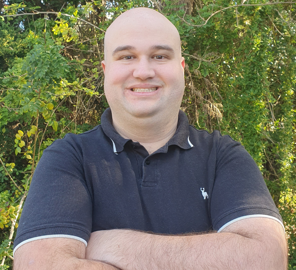 A man with a shaved head smiling with arms crossed in front of a background of green foliage.