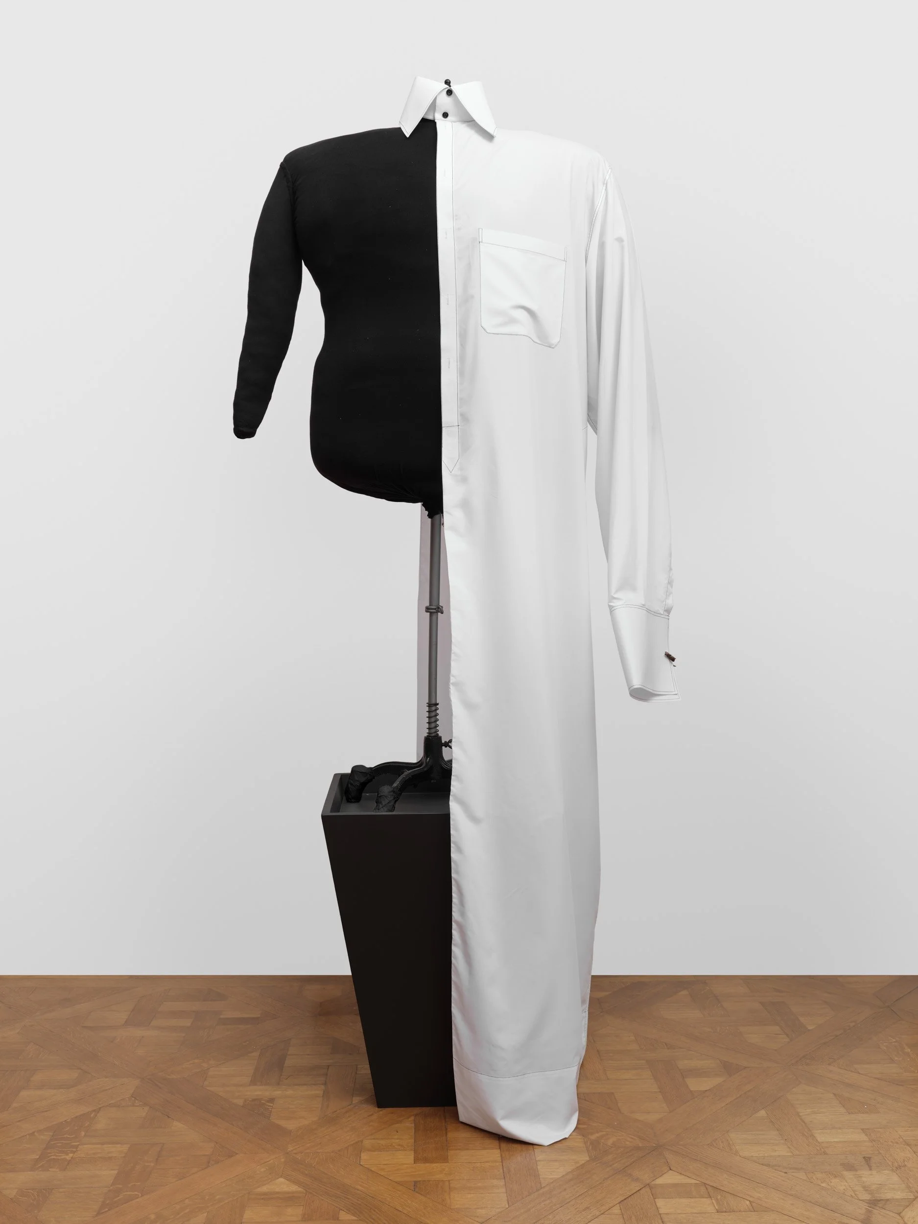 A mannequin with a half black, half white shirt, divided vertically, standing against a plain white background on a wooden floor.
