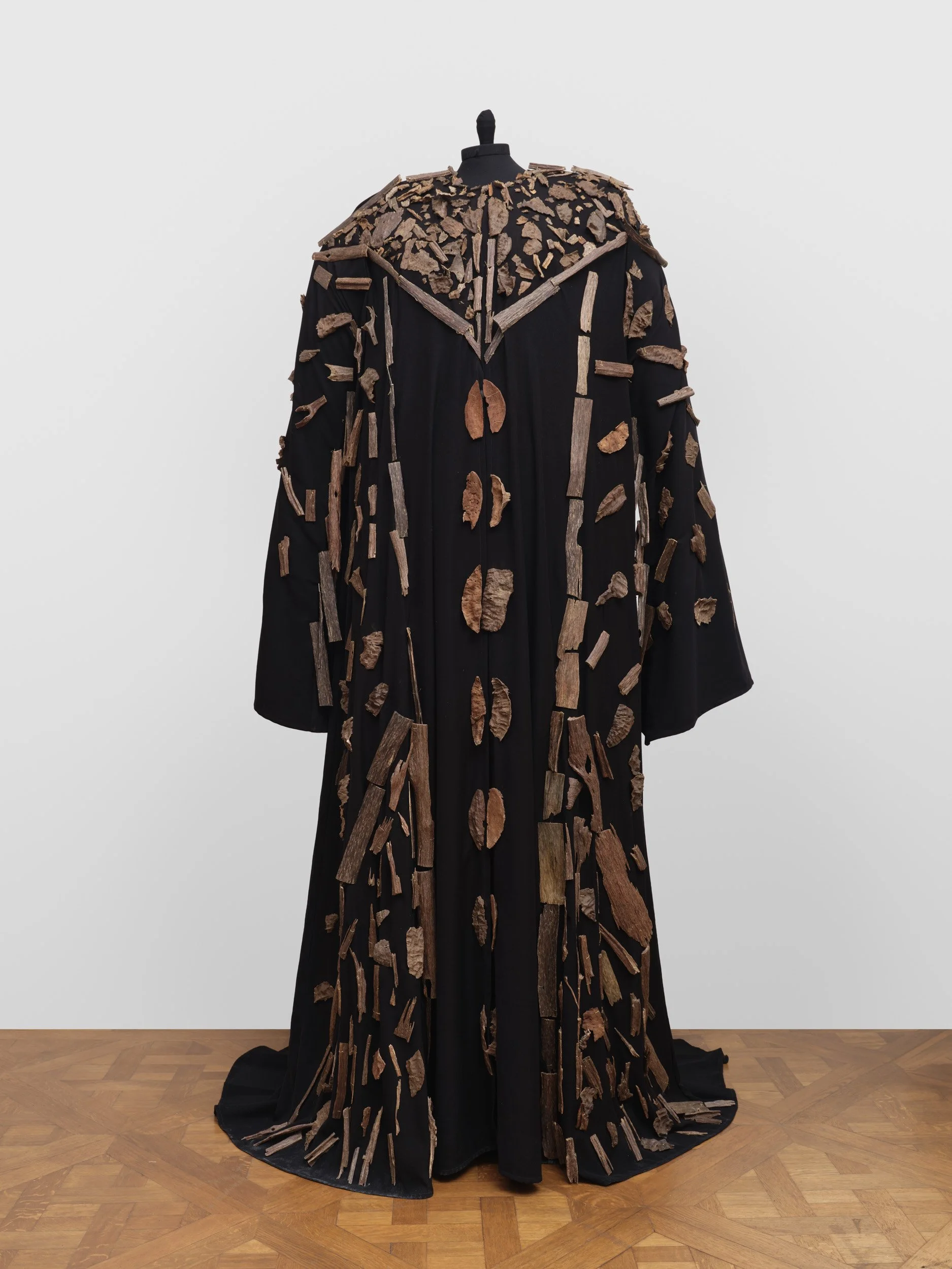 A long black garment displayed on a mannequin, adorned with numerous irregularly shaped wooden fragments attached all over.