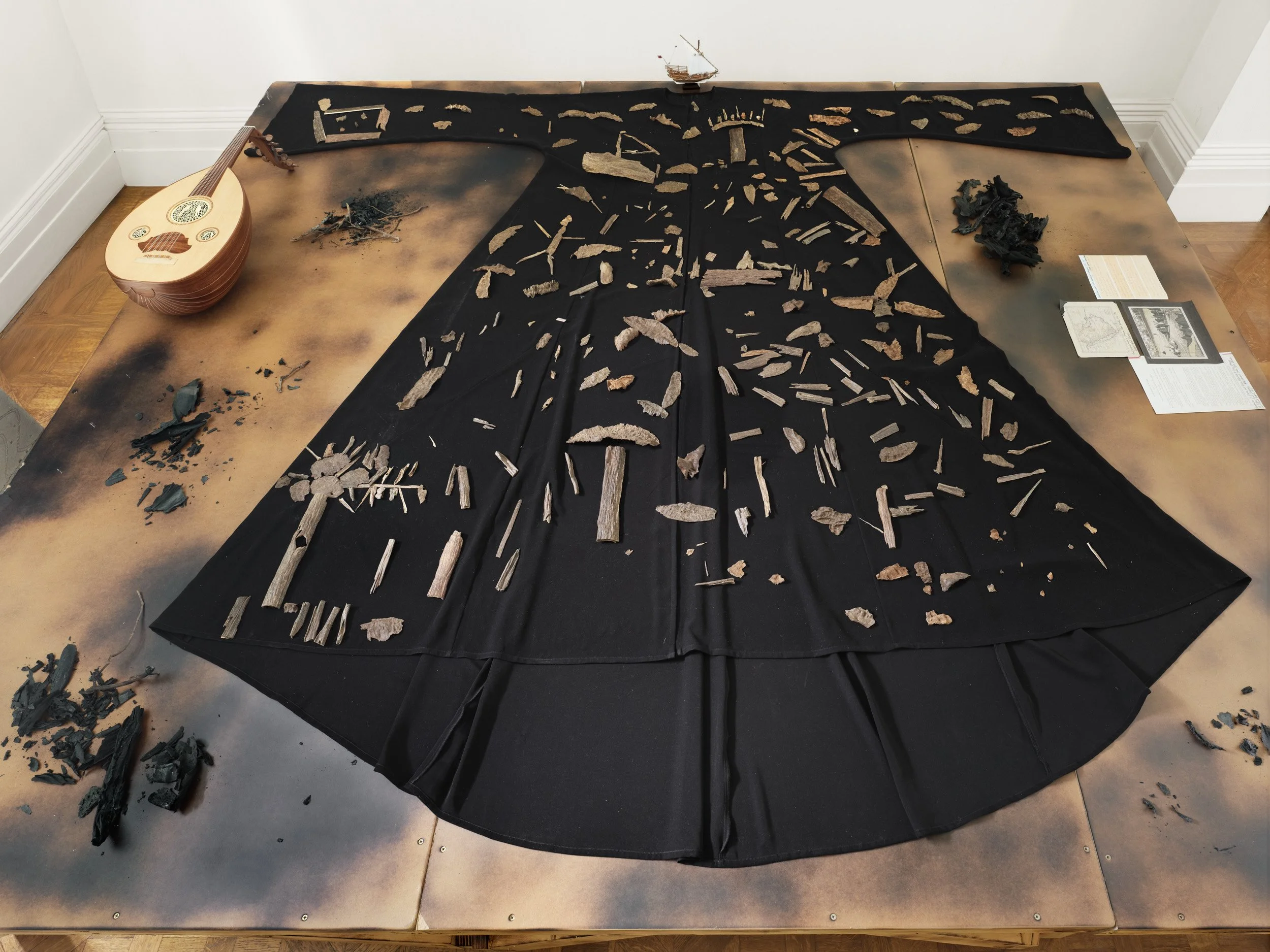 An exhibit of historical artifacts on a table, including a stringed instrument on the left, scattered pottery shards, a small model ship at the back, and informational papers on the right, with a large black cloth covering the table and some artifacts.