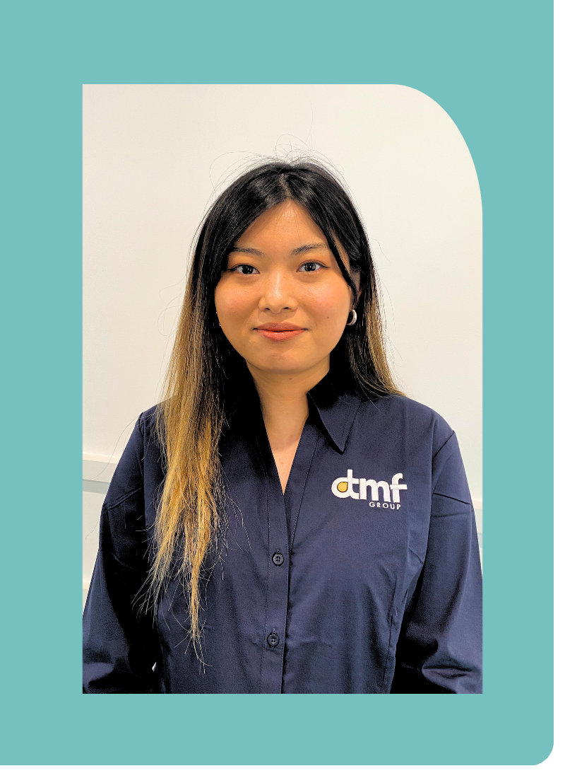 A woman with long dark hair and light brown tips, wearing a navy blue button-up shirt with 'dtmf group' logo, standing against a white wall.