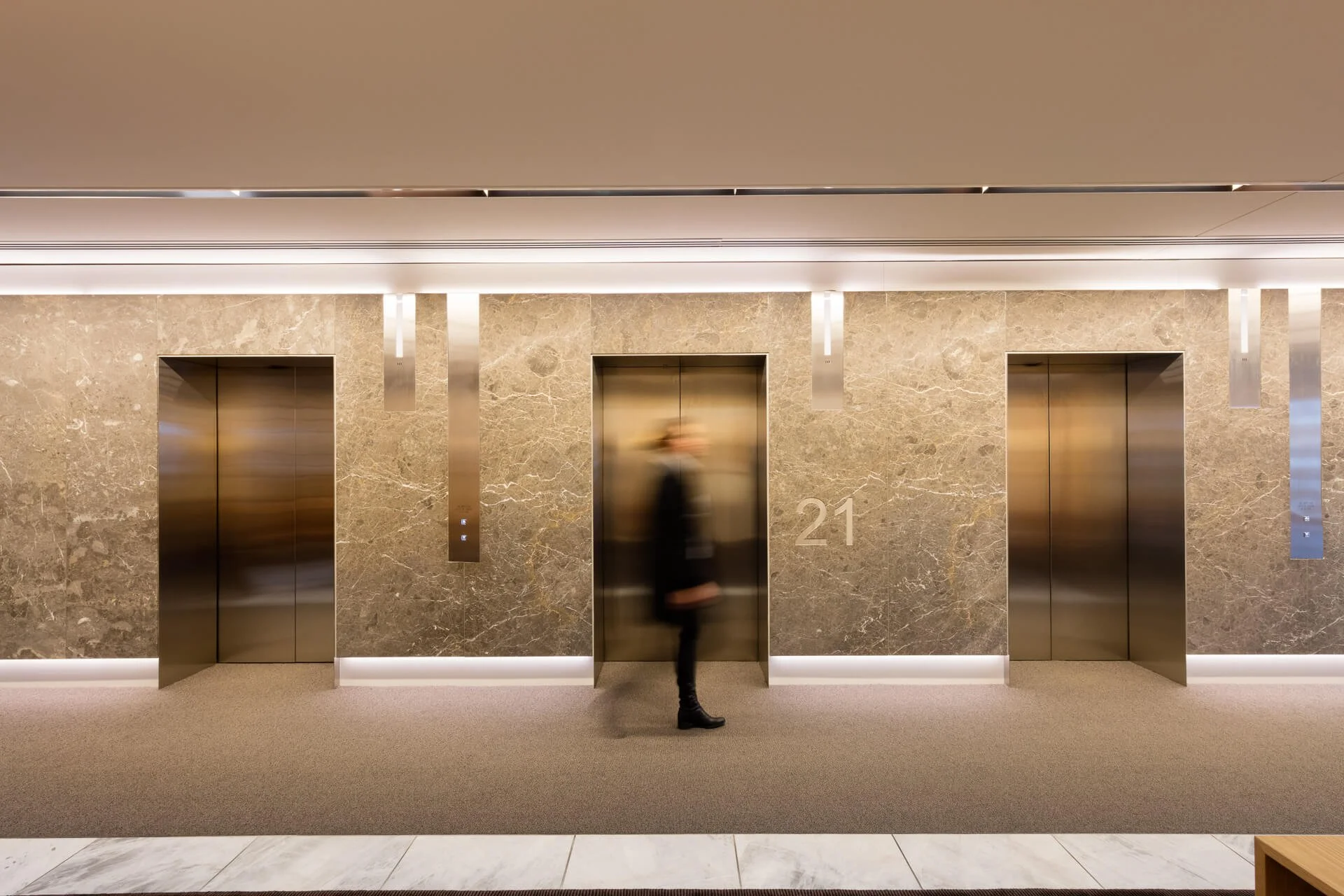 Modern building interior with three elevators, beige marble walls, and a person walking in front of the middle elevator