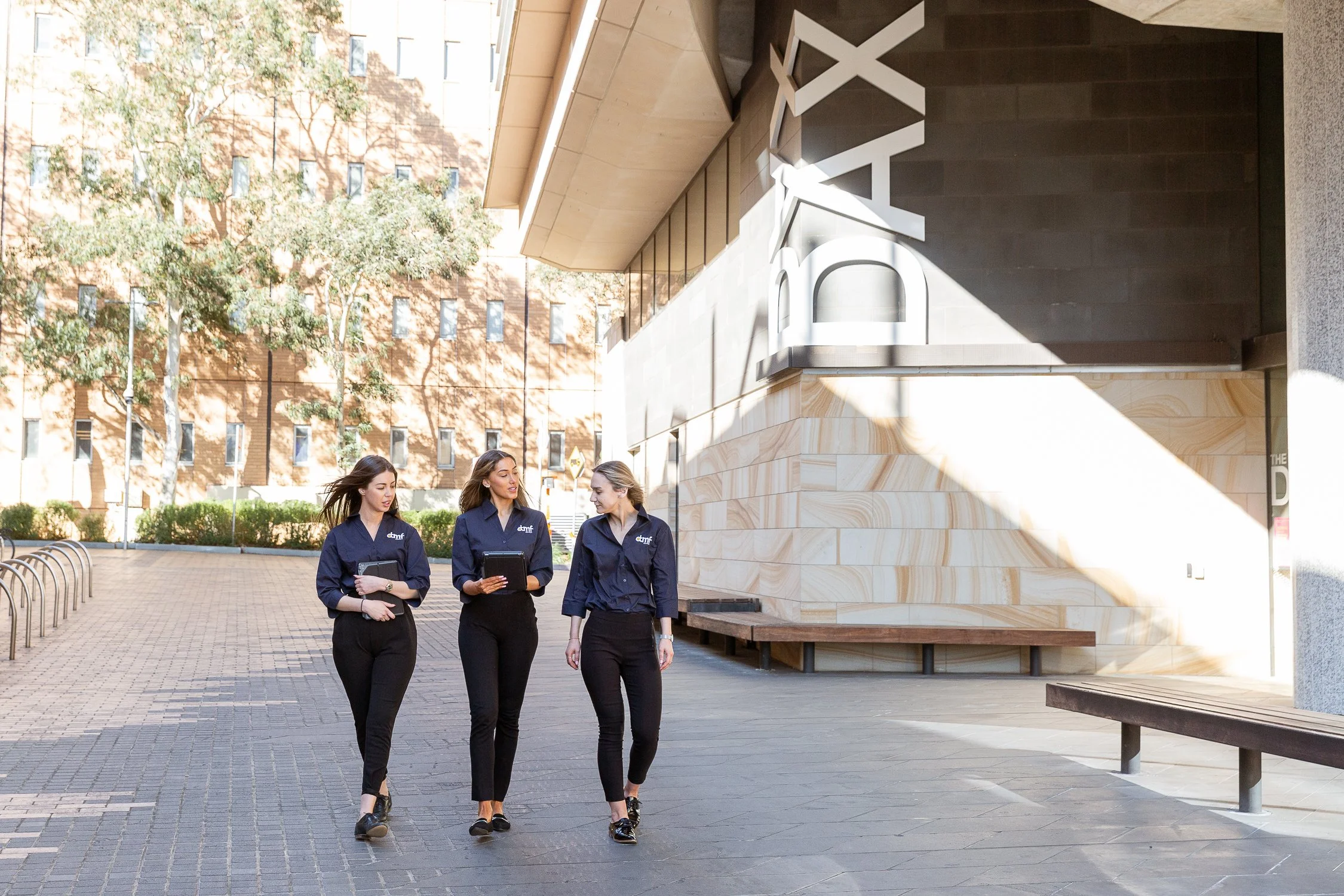 Three women dressed in black business attire with tablet devices walking outdoors on a brick-paved walkway near a modern building with large signs and trees in the background.