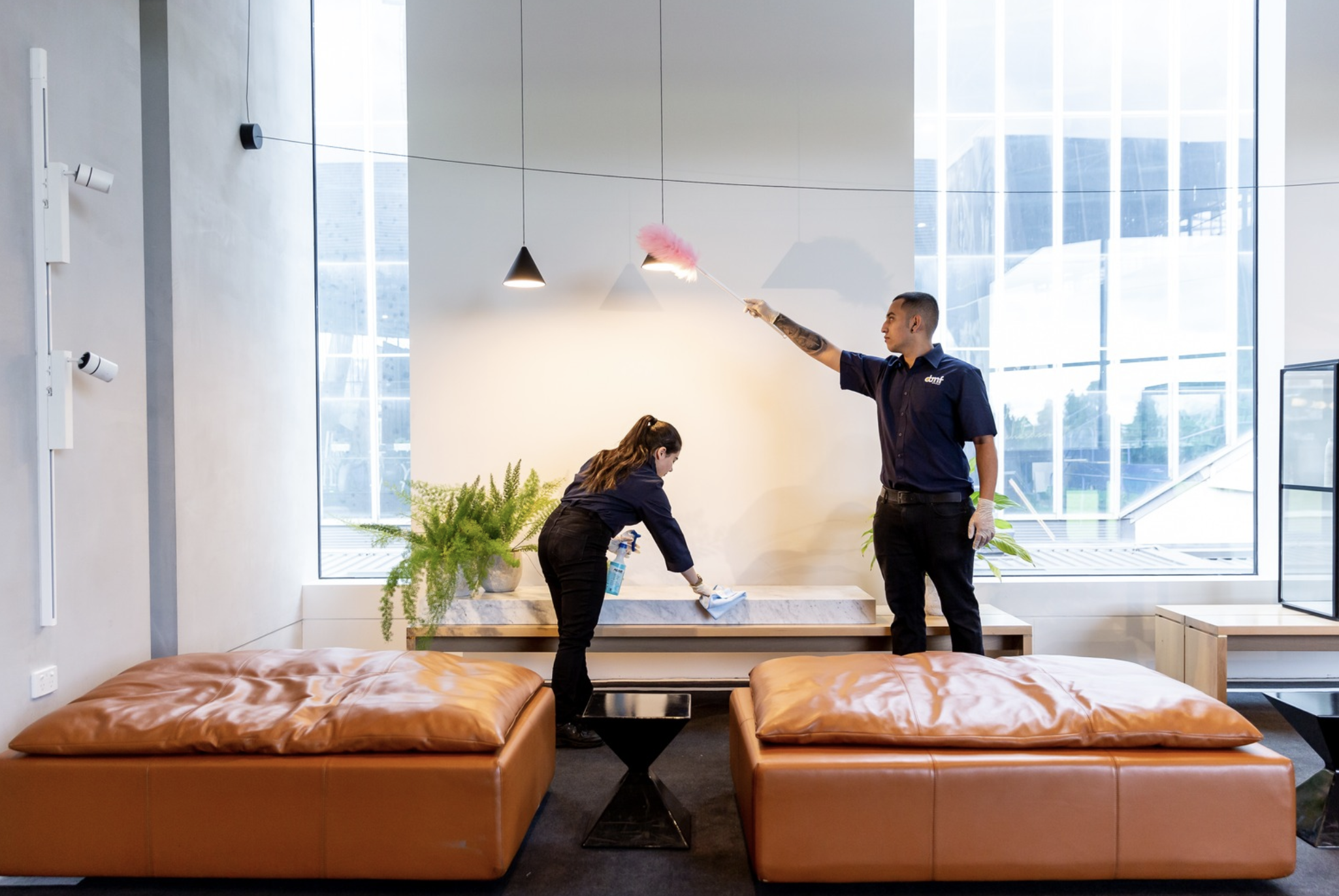 Two workers cleaning a lobby area with plants, large windows, modern lighting, and brown leather couches.