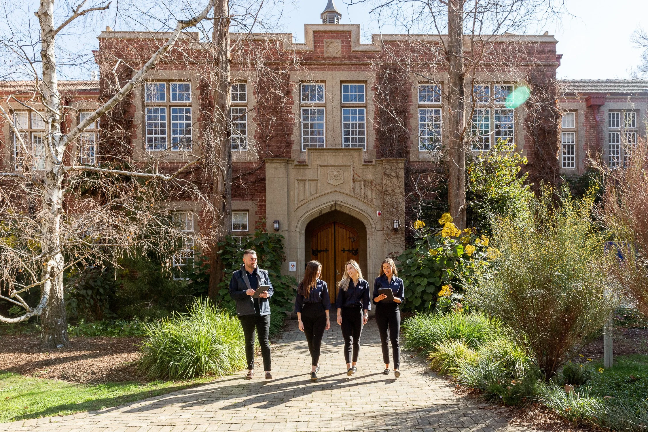 Four young people, two men and two women, walking and talking outside near a large historic brick building with large windows, surrounded by trees and greenery.