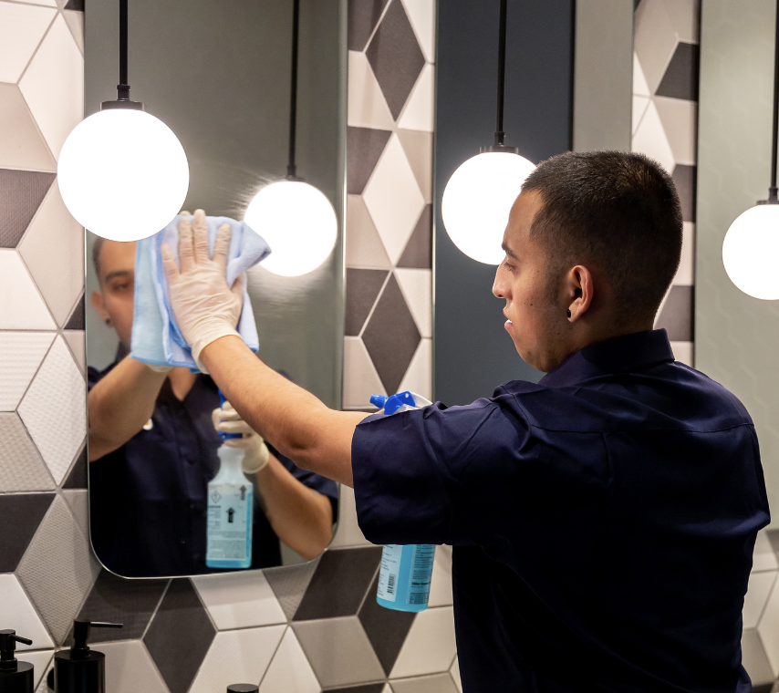 A man cleaning a mirror in a modern bathroom with hexagon tiles and hanging lights.