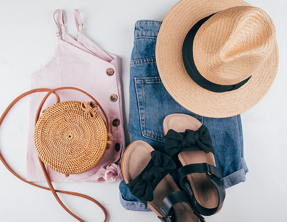 Flat lay of travel accessories including a pink tank top, a woven round handbag, denim shorts, a straw sun hat with a black band, and black sandals with bows.