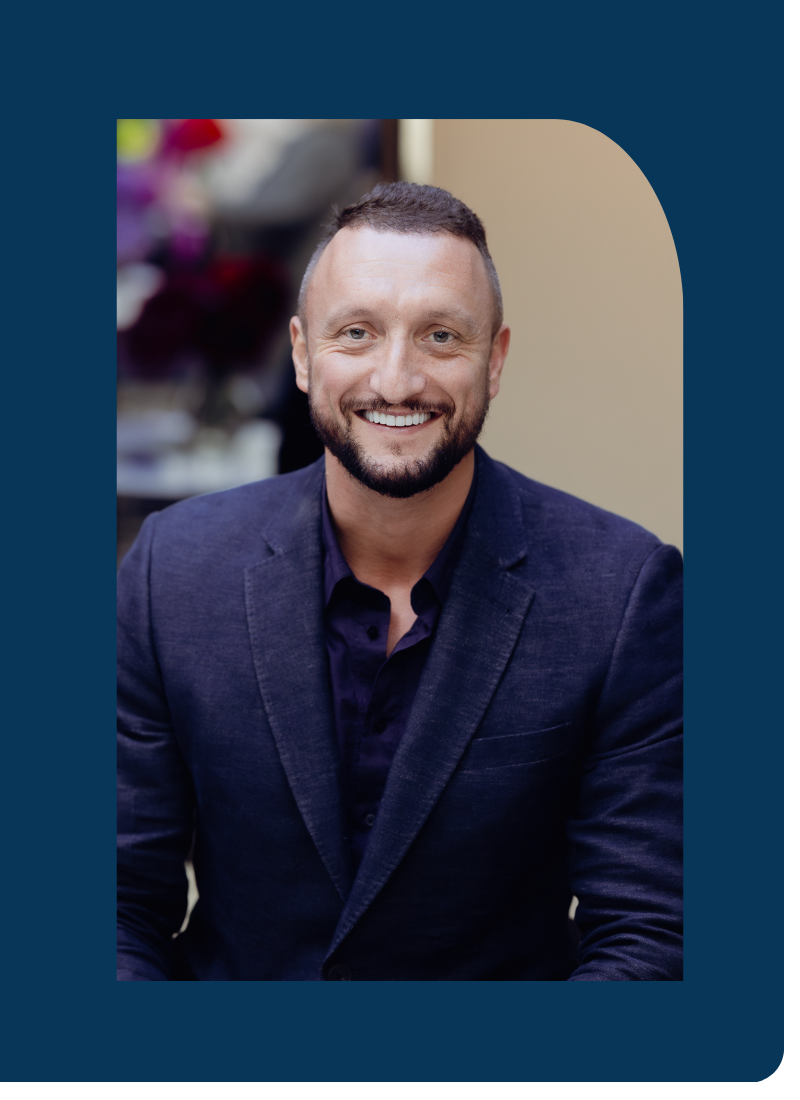 Smiling man with a beard in a dark blazer and dark shirt sitting indoors.
