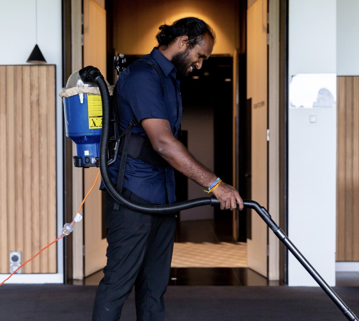 A man with dark curly hair and a beard is wearing a navy blue shirt and black pants while operating a backpack vacuum cleaner to clean the floor indoors.