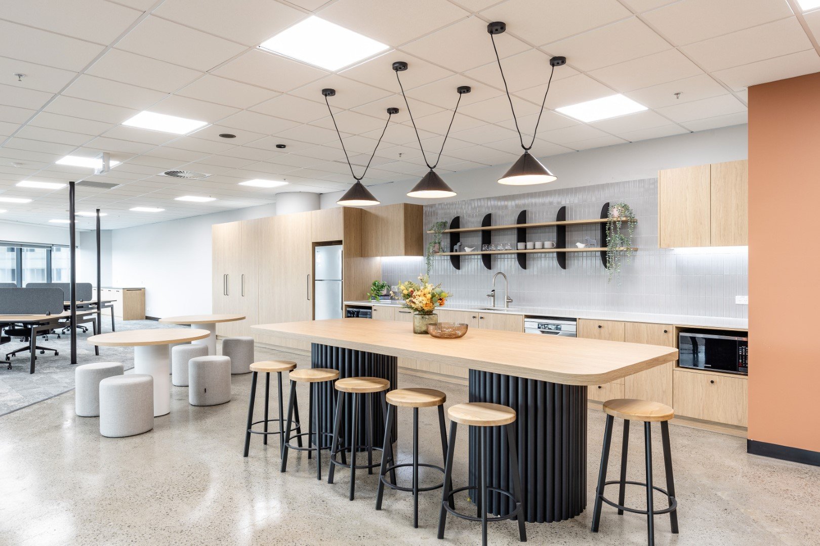 Modern office kitchen with a large wooden island, barstools, hanging pendant lights, open shelves, and a seating area with small round stools and tables.