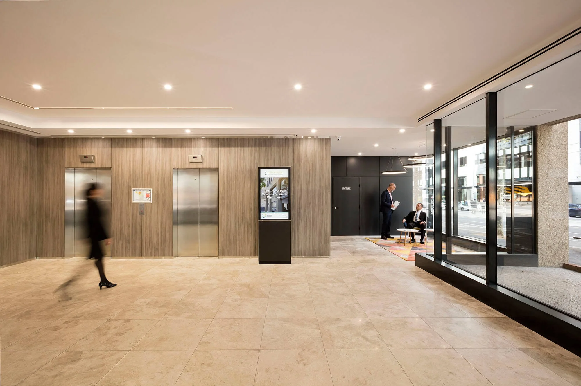Modern lobby with wood-paneled elevator wall, digital information kiosk, and seating area near glass entrance doors. Two men are conversing, and a blurred woman walks past.