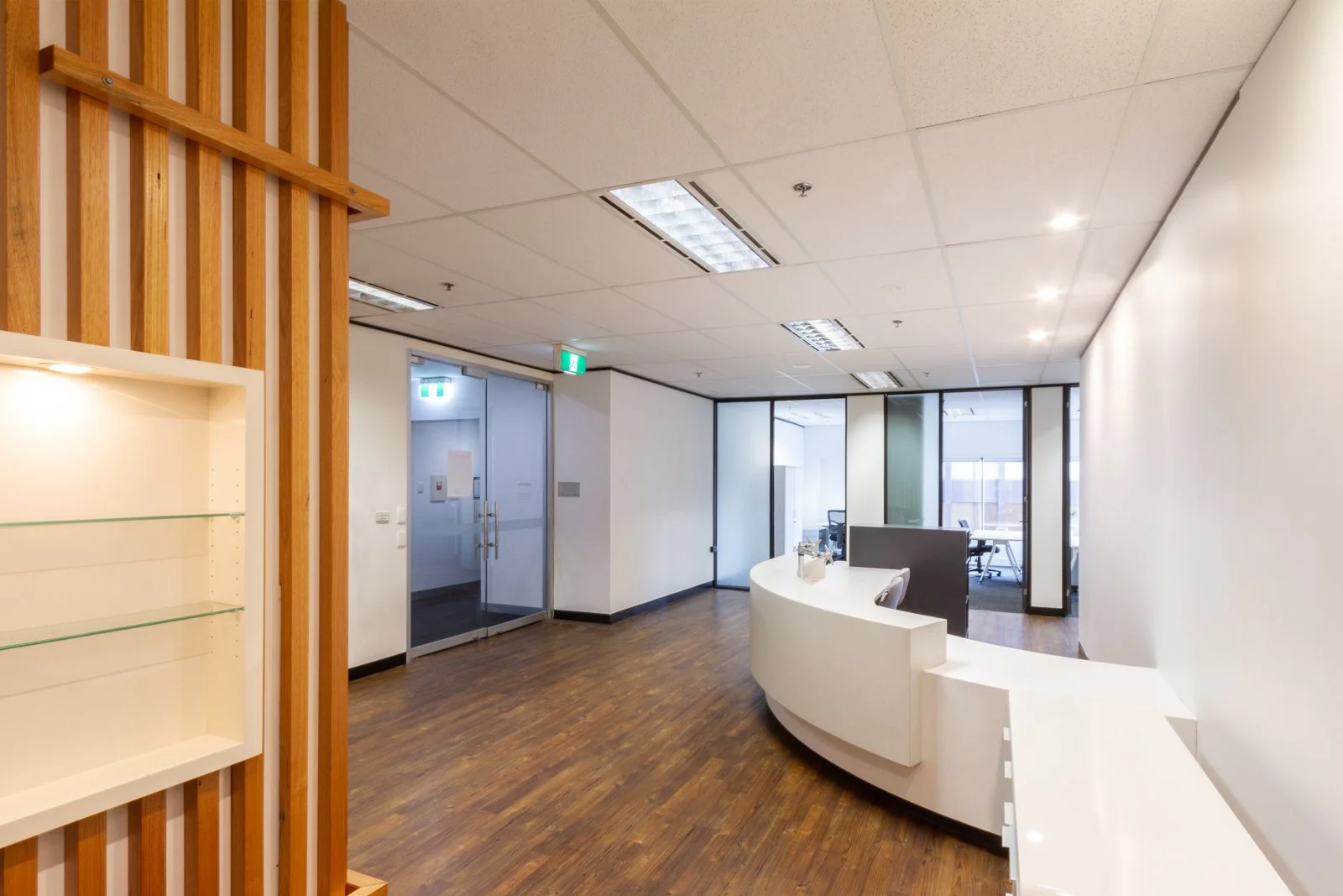 Empty office reception area with a curved white desk, glass walls dividing rooms, and wood flooring.