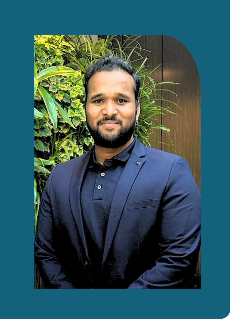 Portrait of a man in a dark blue suit and black shirt, standing in front of green foliage and wooden wall.