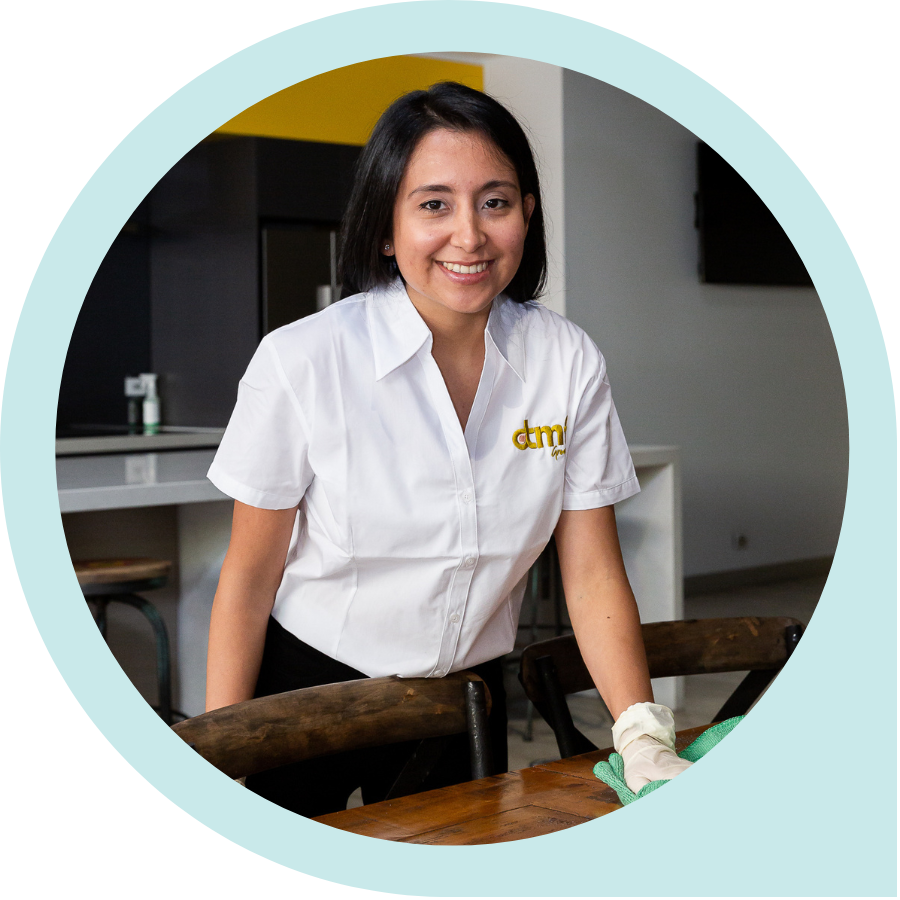 A woman with dark hair smiling, wearing a white shirt with a logo, cleaning a wooden table with a cloth, in a modern kitchen or dining area.