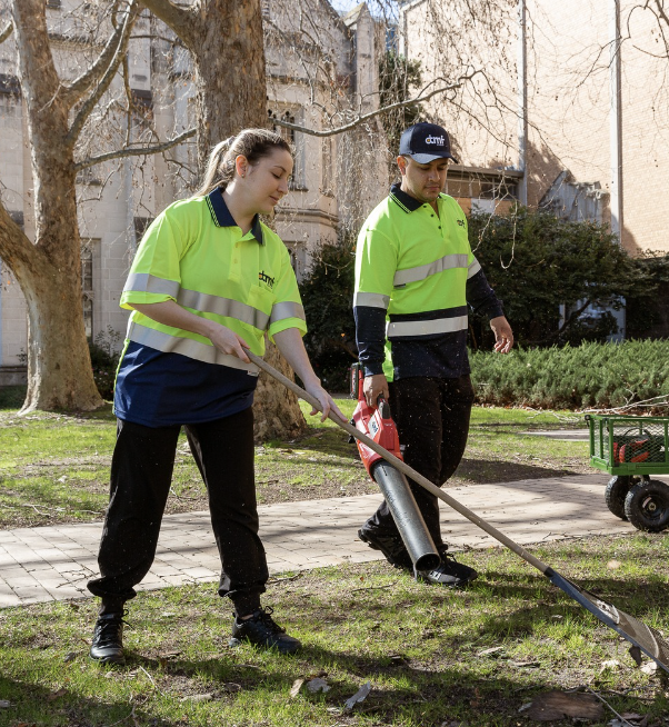 Two workers in high-visibility shirts are working outdoors in a park, one using a digging tool and the other holding a pipe.