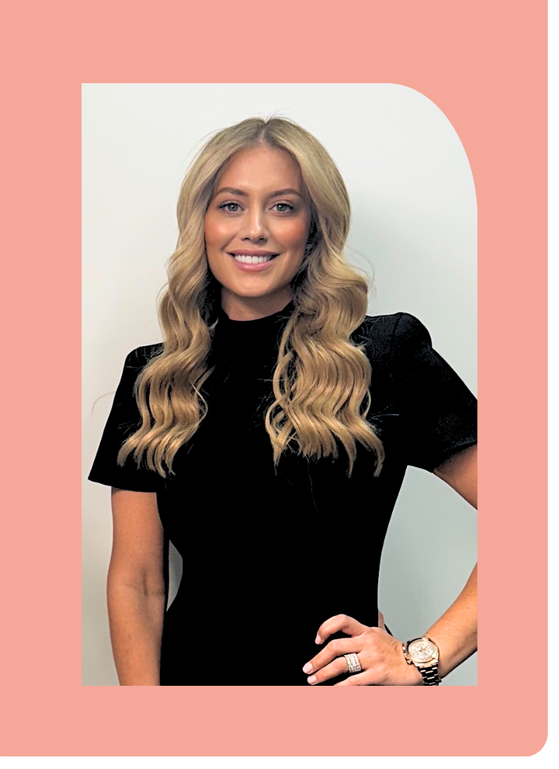 A woman with long blonde hair wearing a black top, smiling, standing against a plain light background.
