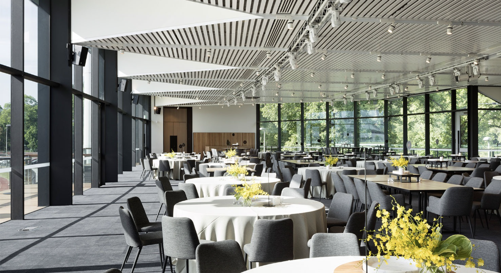 A spacious, modern event space or restaurant with large floor-to-ceiling windows, decorated tables with white tablecloths and yellow floral centerpieces, and a high, patterned ceiling with lighting fixtures.