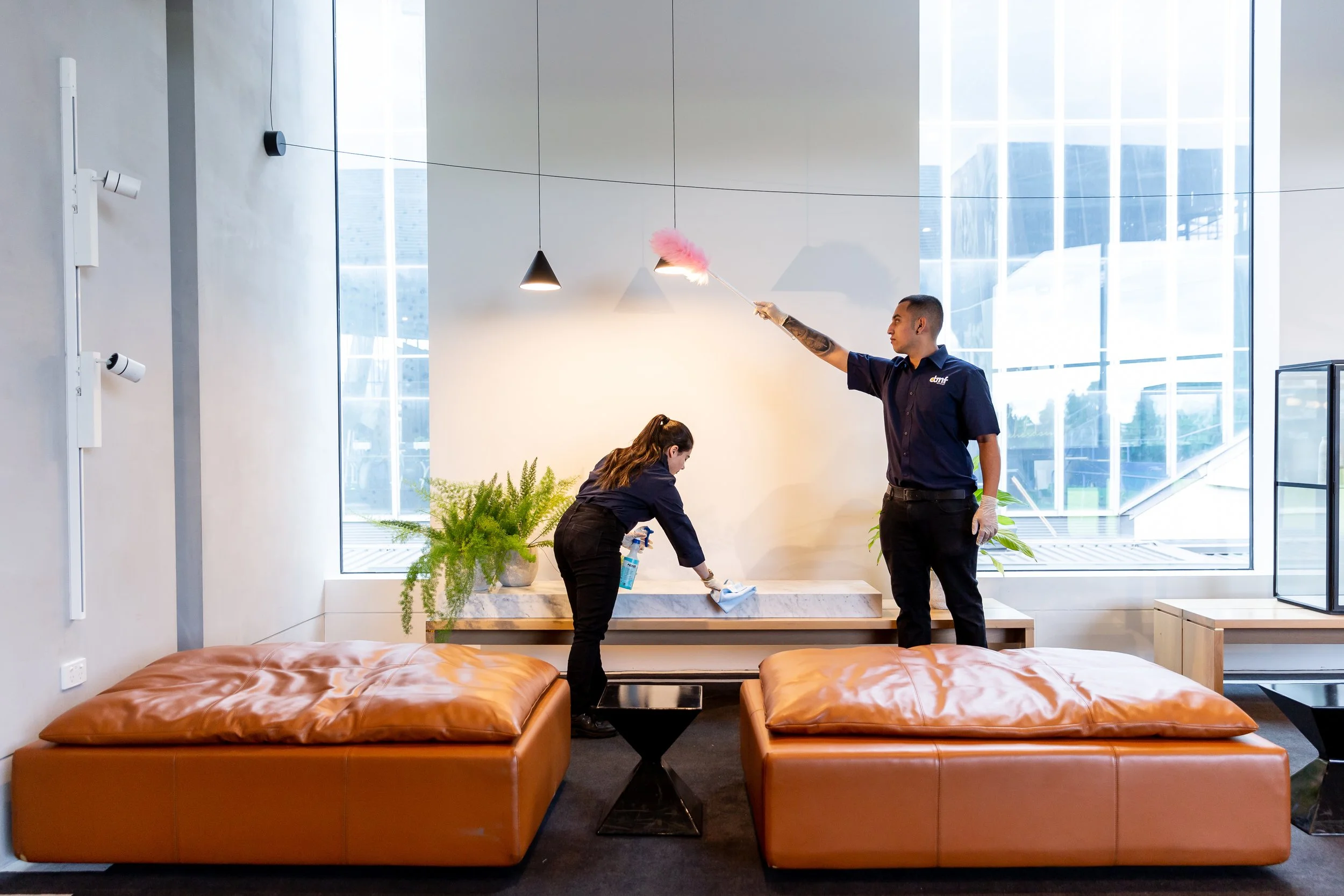 Two individuals cleaning a hotel lobby or lounge area; one man is spraying a cleaning solution onto a surface while holding a pink cleaning tool, and a woman is using a spray bottle and cloth to clean a low surface; there are plants and leather sofas in the foreground, and large windows in the background letting in natural light.