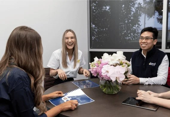 Four people sitting around a table in a meeting room, smiling and laughing, with a large flower arrangement, a tablet, and a brochure on the table.