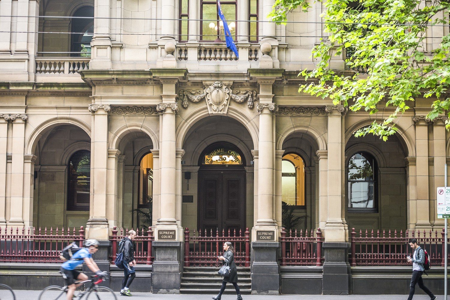 Facade of a historic courthouse building with stone columns and arches, a flag above the entrance, and people walking and biking on the street in front.