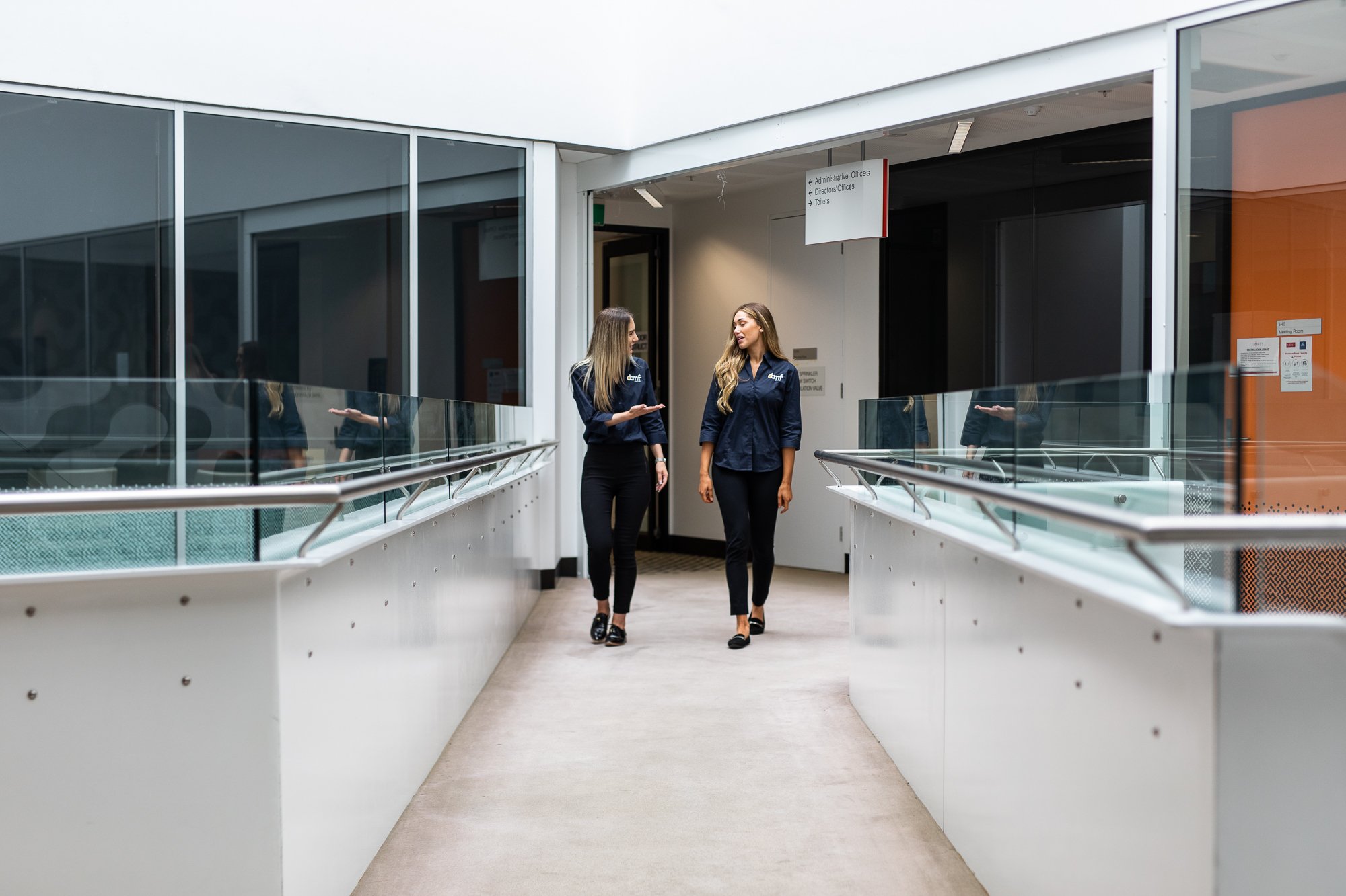 Two women in dark blue shirts walking and talking in a modern indoor public space with glass walls and a railing.
